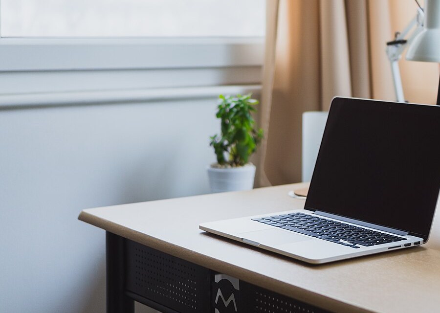 An open silver laptop on a light wooden desk in a bright room