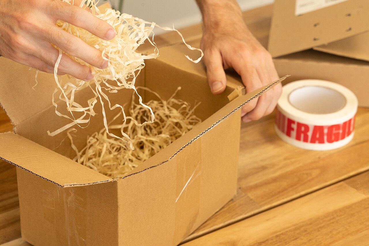Hands packing a cardboard box with shredded paper filler beside a roll of fragile tape
